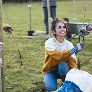 A woman kneels on grass and holds a sapling while participating in an outdoor tree-planting activity with others.