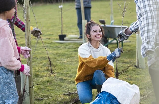 A woman kneels on grass and holds a sapling while participating in an outdoor tree-planting activity with others.