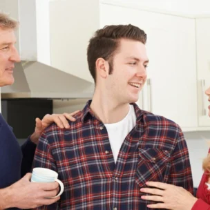 Three people stand in a kitchen having a conversation; an older man holds a mug while a younger man and an older woman face each other and smile.