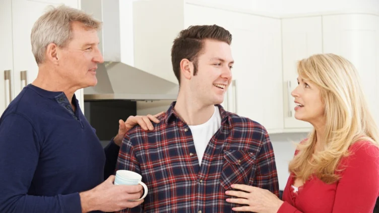 Three people stand in a kitchen having a conversation; an older man holds a mug while a younger man and an older woman face each other and smile.