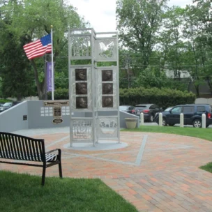 A metal memorial structure with plaques and photos stands on a brick plaza, flanked by benches, with an American flag and parked cars in the background.