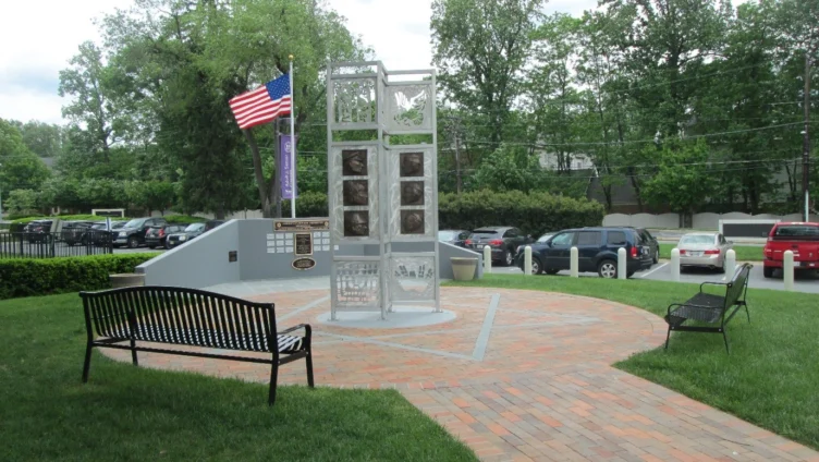 A metal memorial structure with plaques and photos stands on a brick plaza, flanked by benches, with an American flag and parked cars in the background.