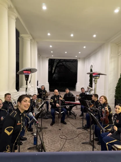 A group of uniformed musicians sits in a semicircle with instruments under a white, columned portico at night. One woman smiles at the camera in the foreground.