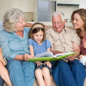 Three adults and three children sit on a couch in a bright living room, smiling and looking at a large book held by the girl in the center.