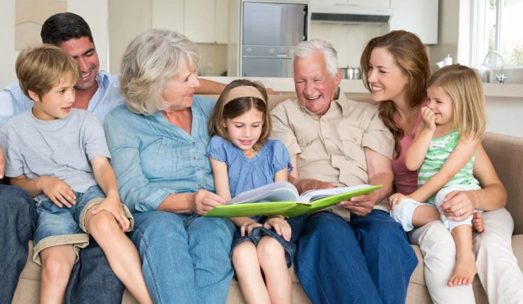 Three adults and three children sit on a couch in a bright living room, smiling and looking at a large book held by the girl in the center.