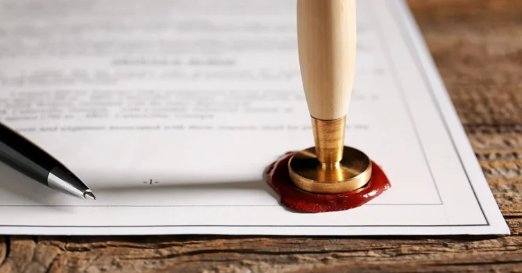 A document on a wooden table is sealed with red wax and a stamp; a black pen lies next to it.
