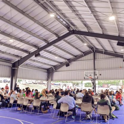 A large group of people sit at tables under a covered outdoor basketball court, participating in an organized event or meeting.