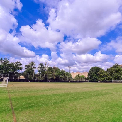 A large, empty soccer field with two goals, surrounded by trees and buildings, under a blue sky with scattered clouds.