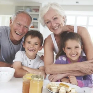An older couple smiles with two young children at a kitchen table set with breakfast foods such as fruit, jars, and bowls.