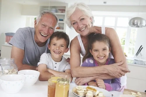 An older couple smiles with two young children at a kitchen table set with breakfast foods such as fruit, jars, and bowls.