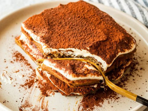 A square piece of tiramisu with cocoa powder on top, partially eaten with a gold fork resting on a white plate.