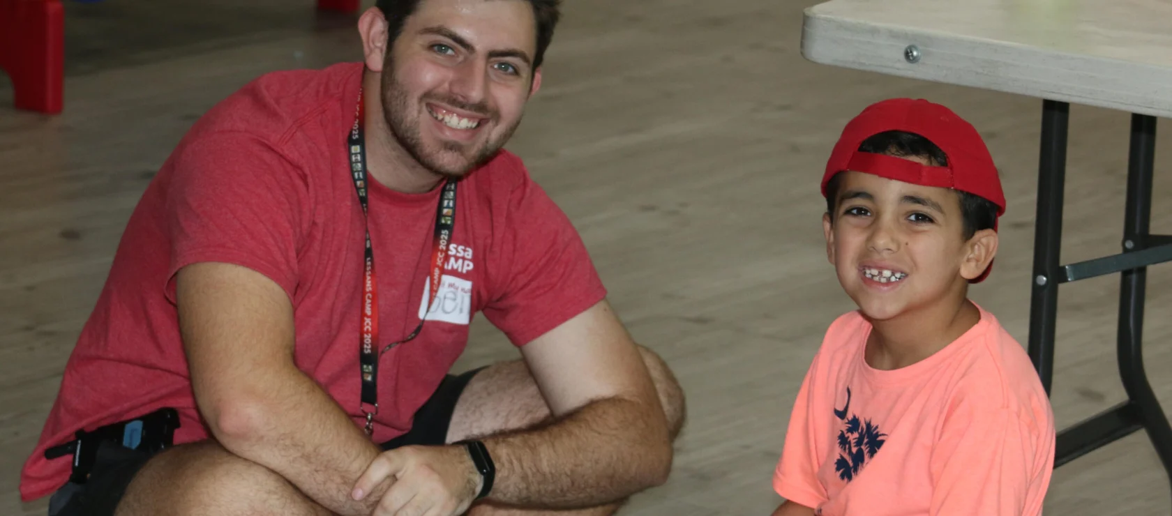An adult and a child, both smiling, sit on the floor next to a table in a recreational indoor setting.