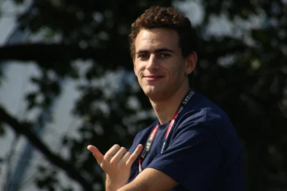 A young man wearing a navy blue shirt and lanyard smiles and makes the shaka hand gesture with trees in the blurred background.