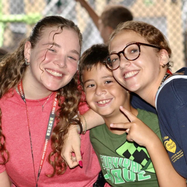 Three people pose and smile at the camera; two young women on either side of a young boy who is seated between them, all in casual clothes with a chain-link fence in the background.