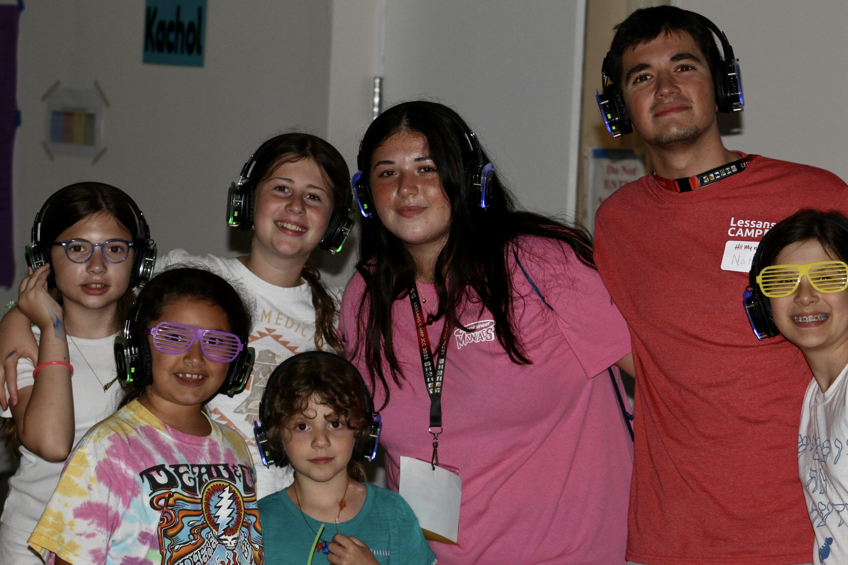 A group of children and two adults wearing wireless headphones and colorful glasses pose together indoors, smiling at the camera.