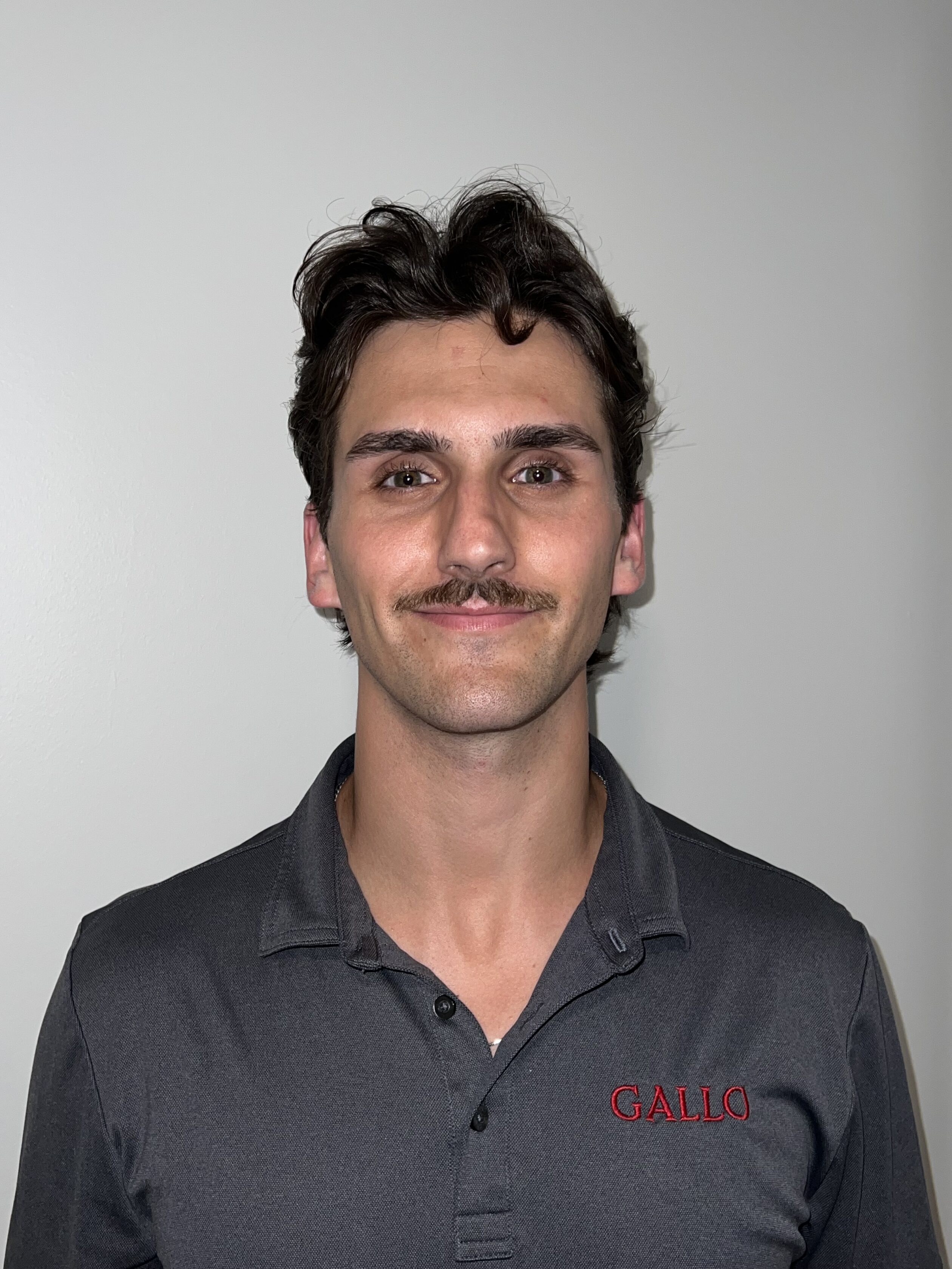 A man with dark hair and a mustache wears a dark gray collared shirt with "GALLO" embroidered in red, standing against a plain light-colored wall.