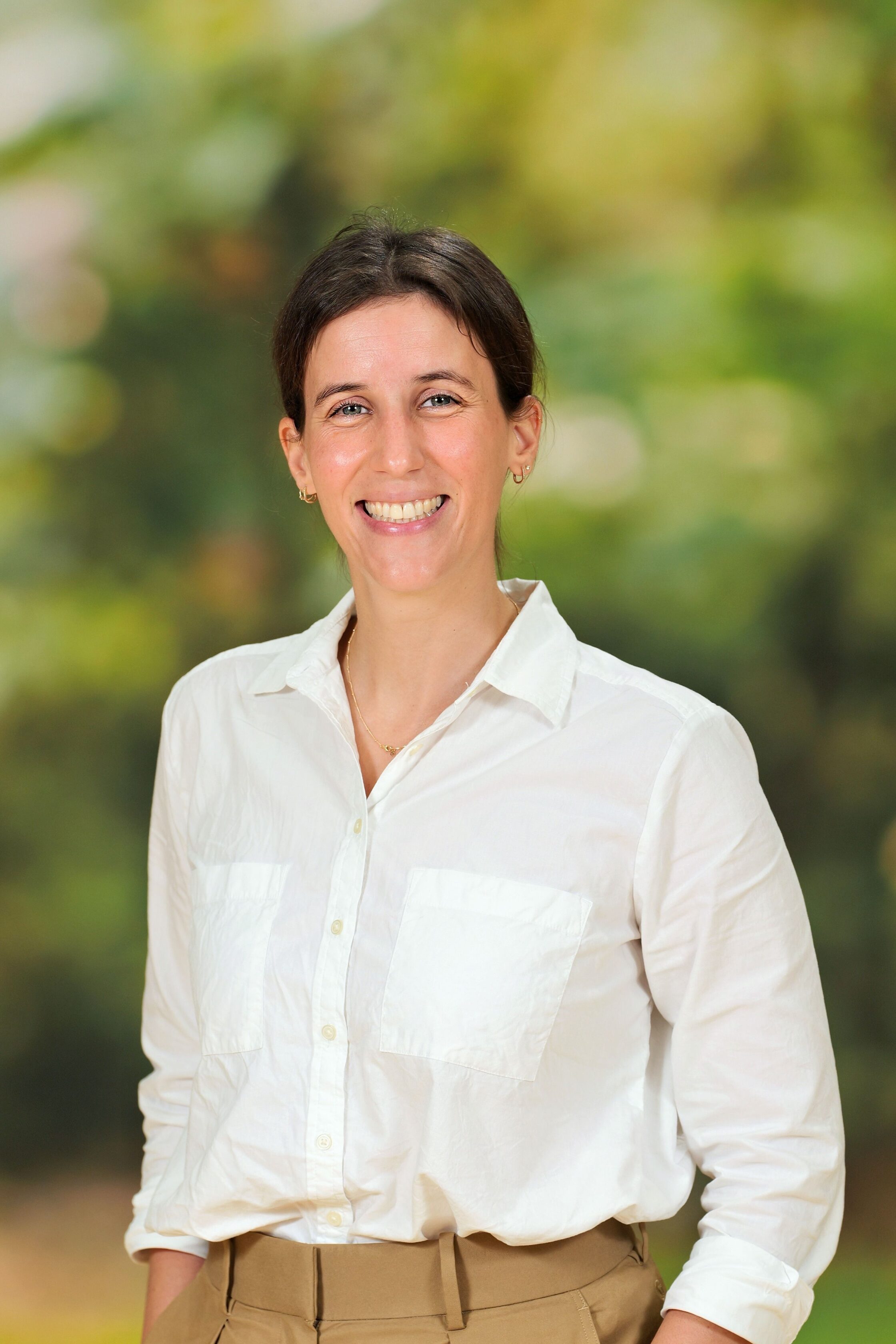 A woman with dark hair tied back, wearing a white button-up shirt and beige pants, stands smiling in front of a blurred outdoor background.