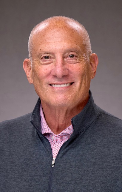Smiling older man with short gray hair wearing a dark zip-up jacket and a light pink collared shirt, posed against a plain gray background.