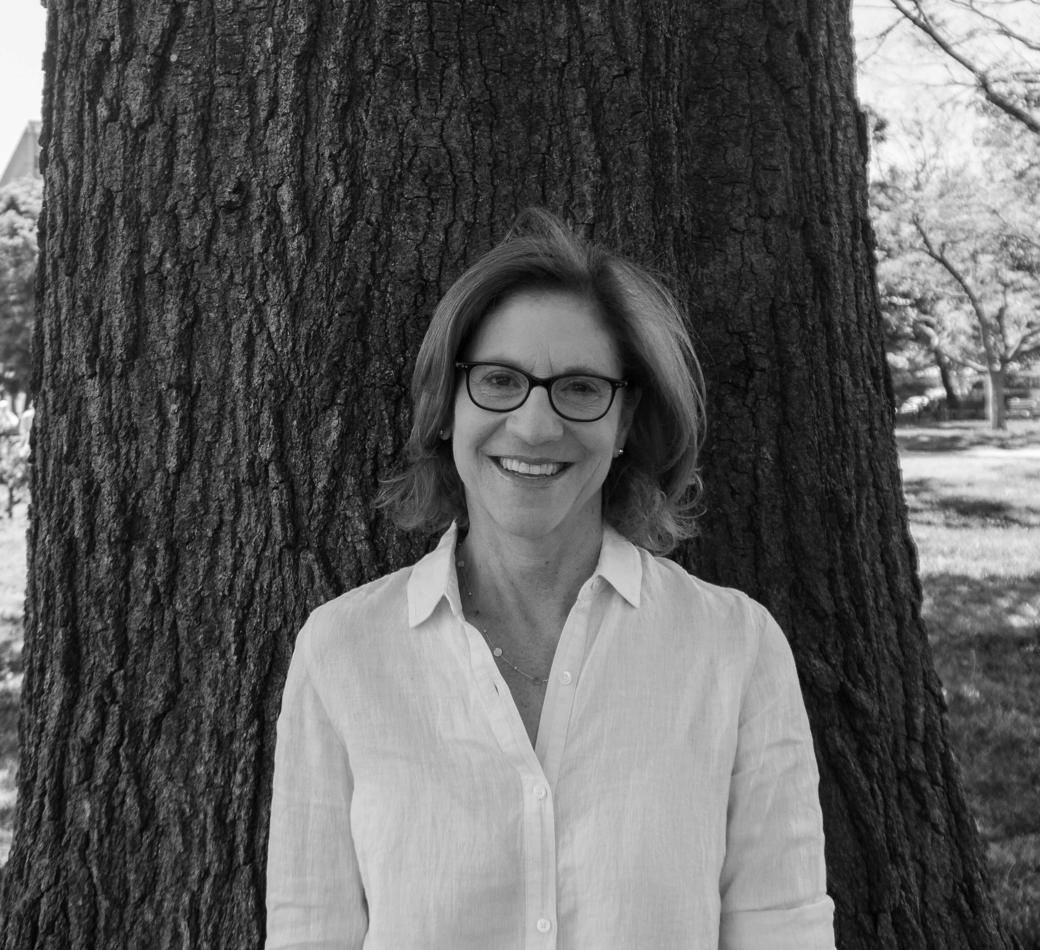 A woman wearing glasses and a light-colored blouse stands in front of a large tree, smiling. The photo is in black and white.