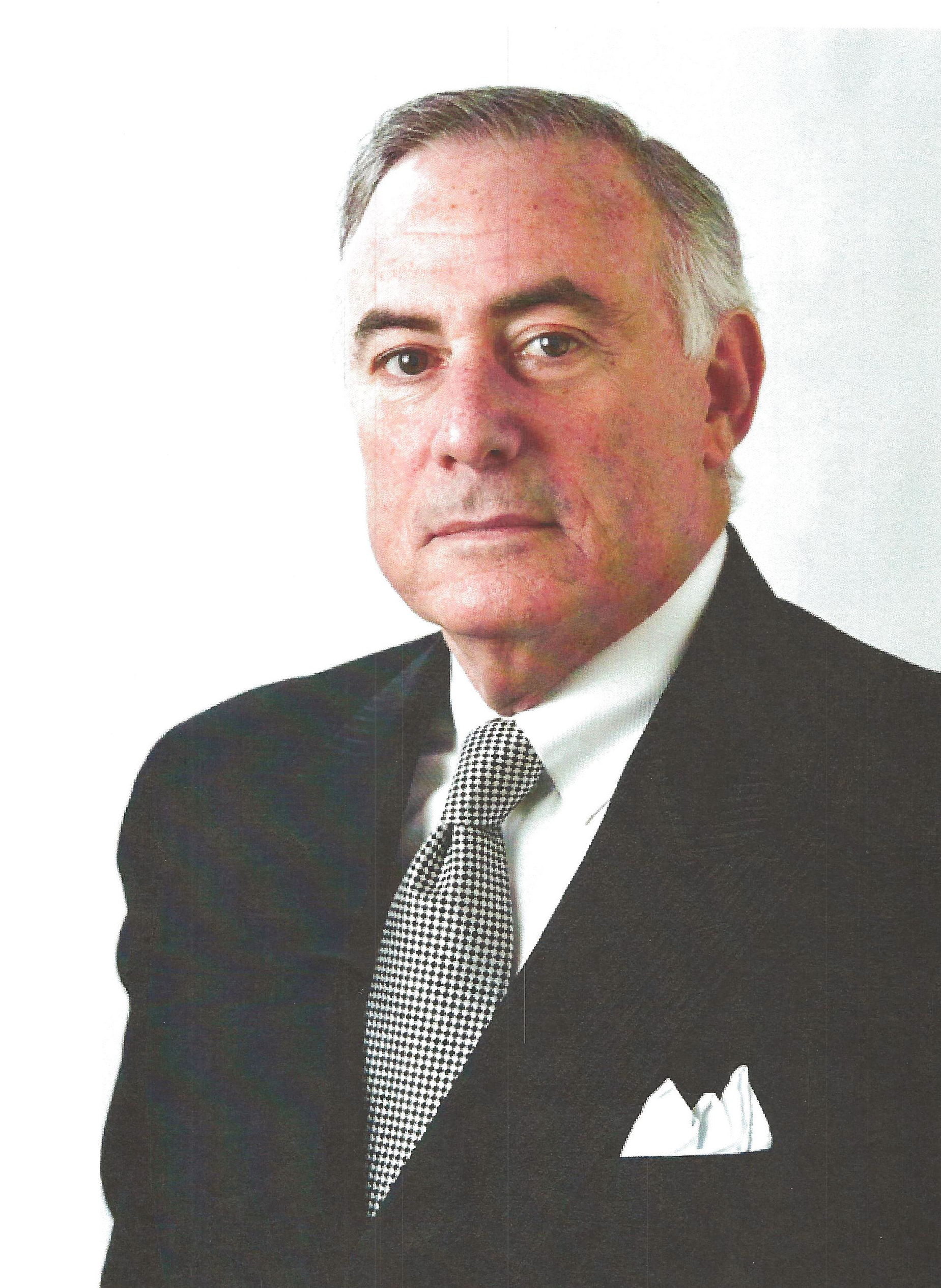A mature man with gray hair wearing a dark suit, patterned tie, and white pocket square stands against a plain white background, looking directly at the camera.