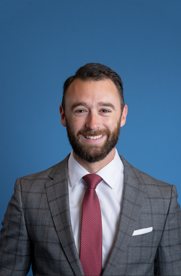 A man with a beard wearing a gray checked suit, white shirt, and red tie stands in front of a plain blue background, smiling at the camera.