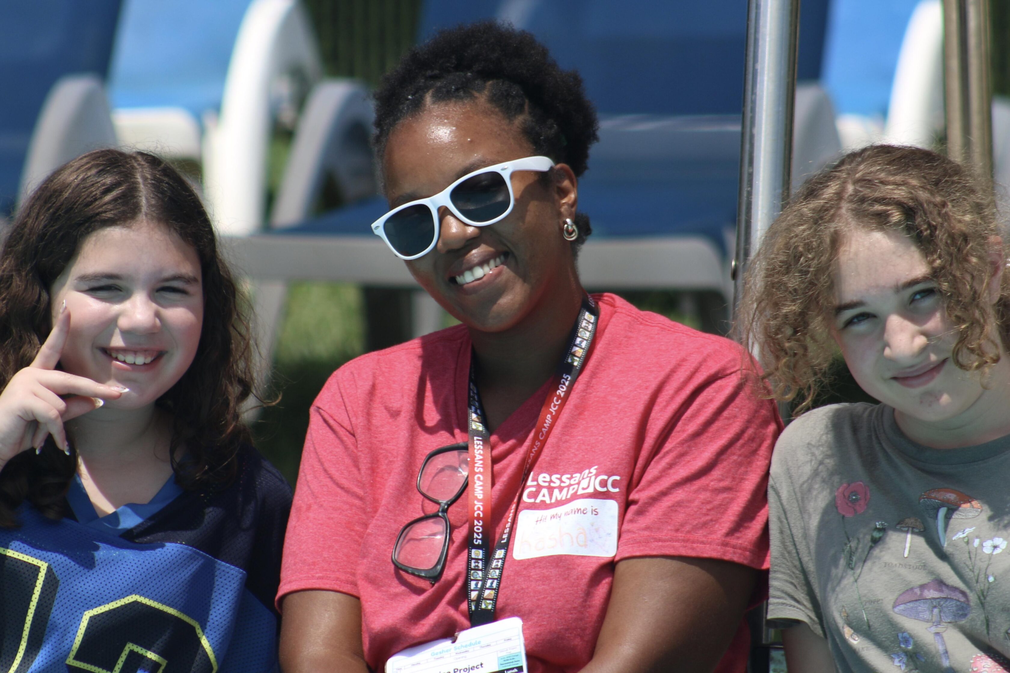 Three people sit outdoors on lounge chairs. The person in the center wears sunglasses and a red shirt with a name tag, flanked by two smiling individuals.
