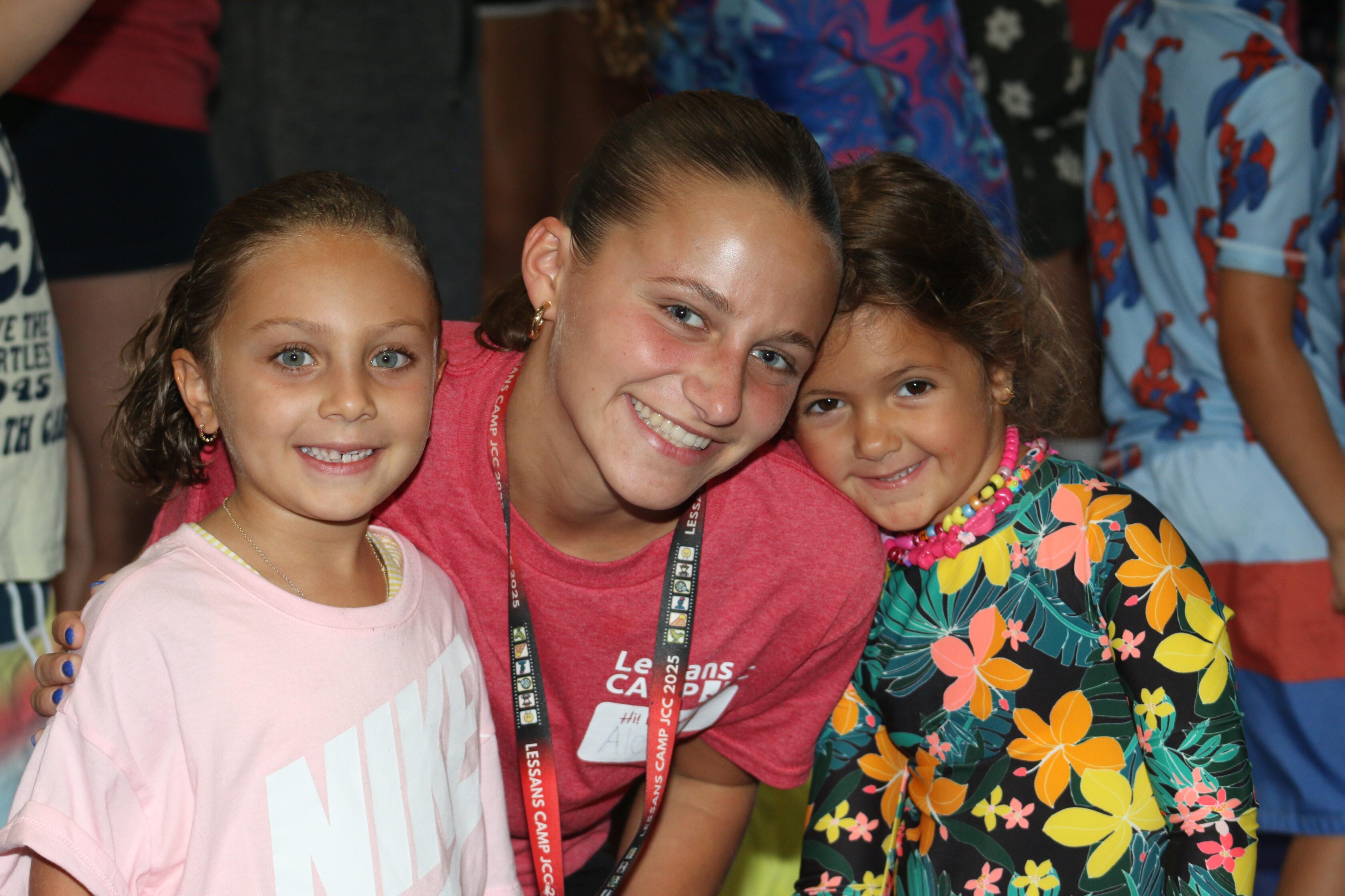 A young woman poses and smiles with two young girls, all facing the camera. The girls wear casual clothes, and other people are visible in the background.
