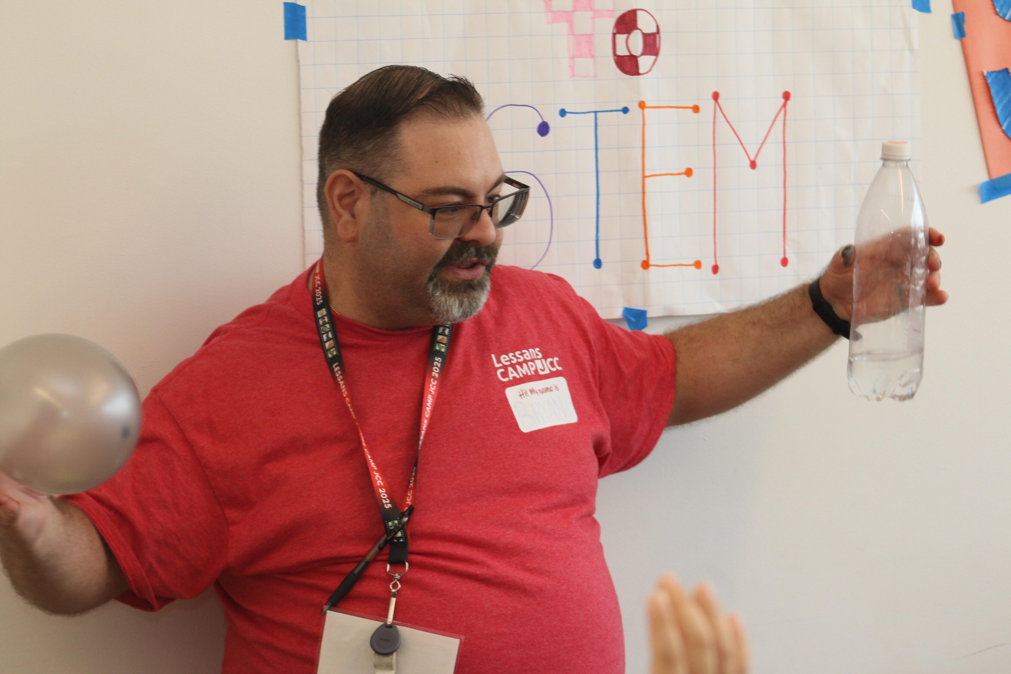 A man in a red shirt holds a balloon and a plastic bottle in front of a wall sign that reads "STEM" with colorful decorations.