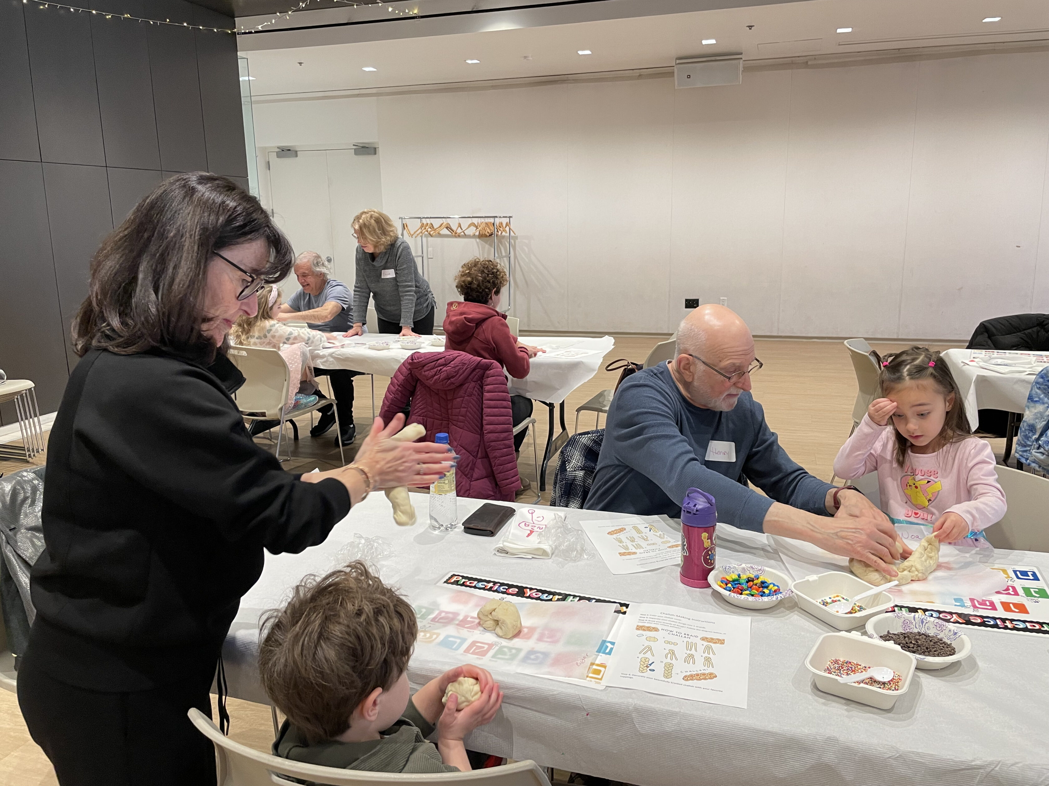 Adults and children sit at tables working with dough and colorful toppings, participating in a hands-on activity in a spacious, well-lit room.