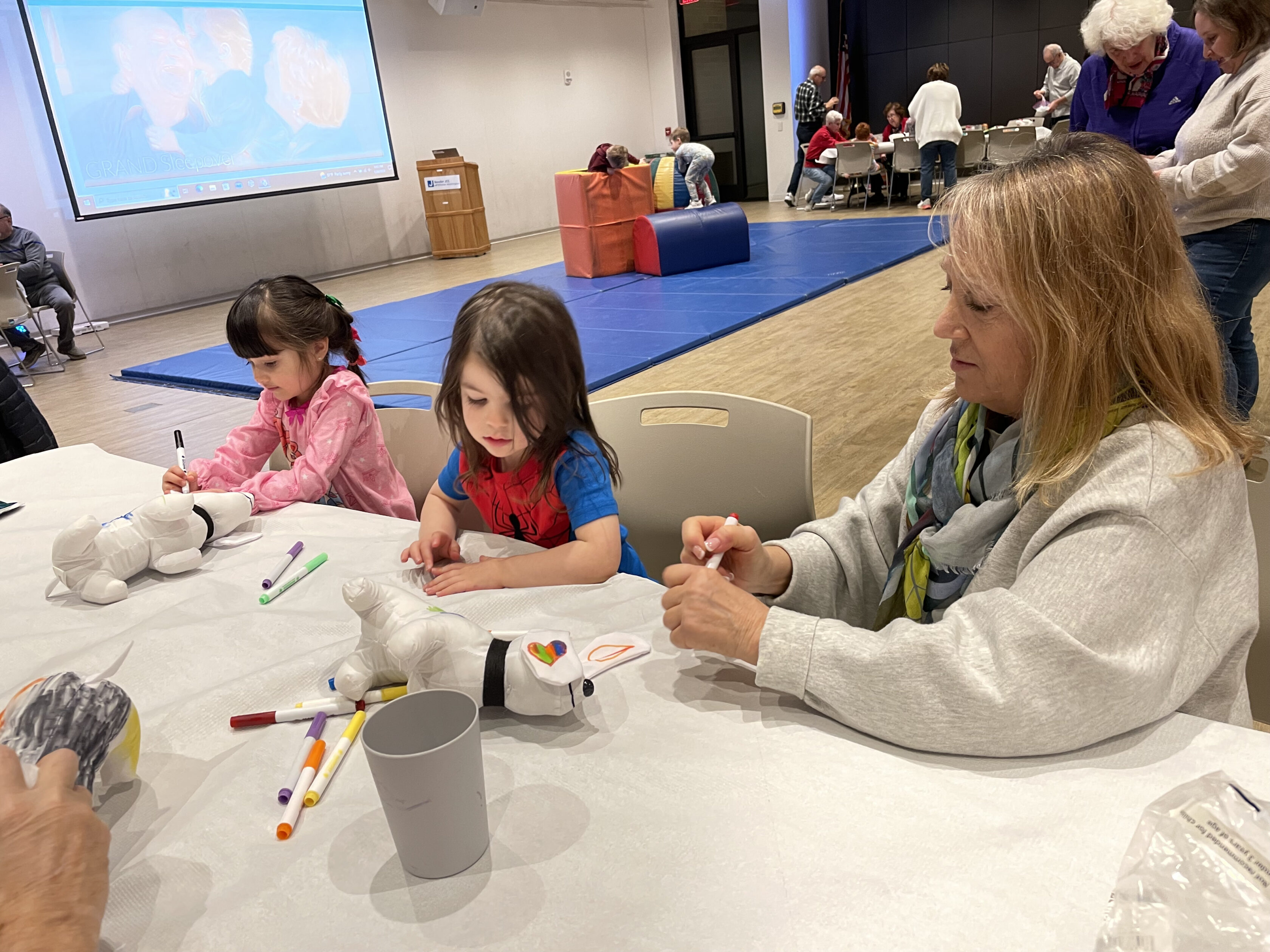 Two young girls and an adult woman sit at a table coloring stuffed animal toys with markers in a spacious, well-lit room with other people in the background.