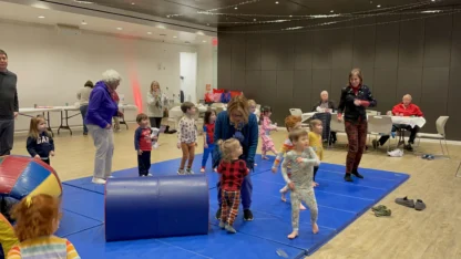 Children in pajamas play and jump on blue mats in a community room, supervised by several adults; some people sit at tables in the background.