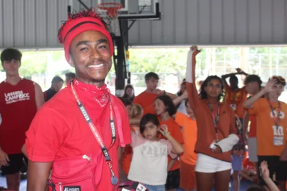A young person in a red outfit smiles at the camera while a group of children and teens in orange shirts gather in the background inside a gymnasium.