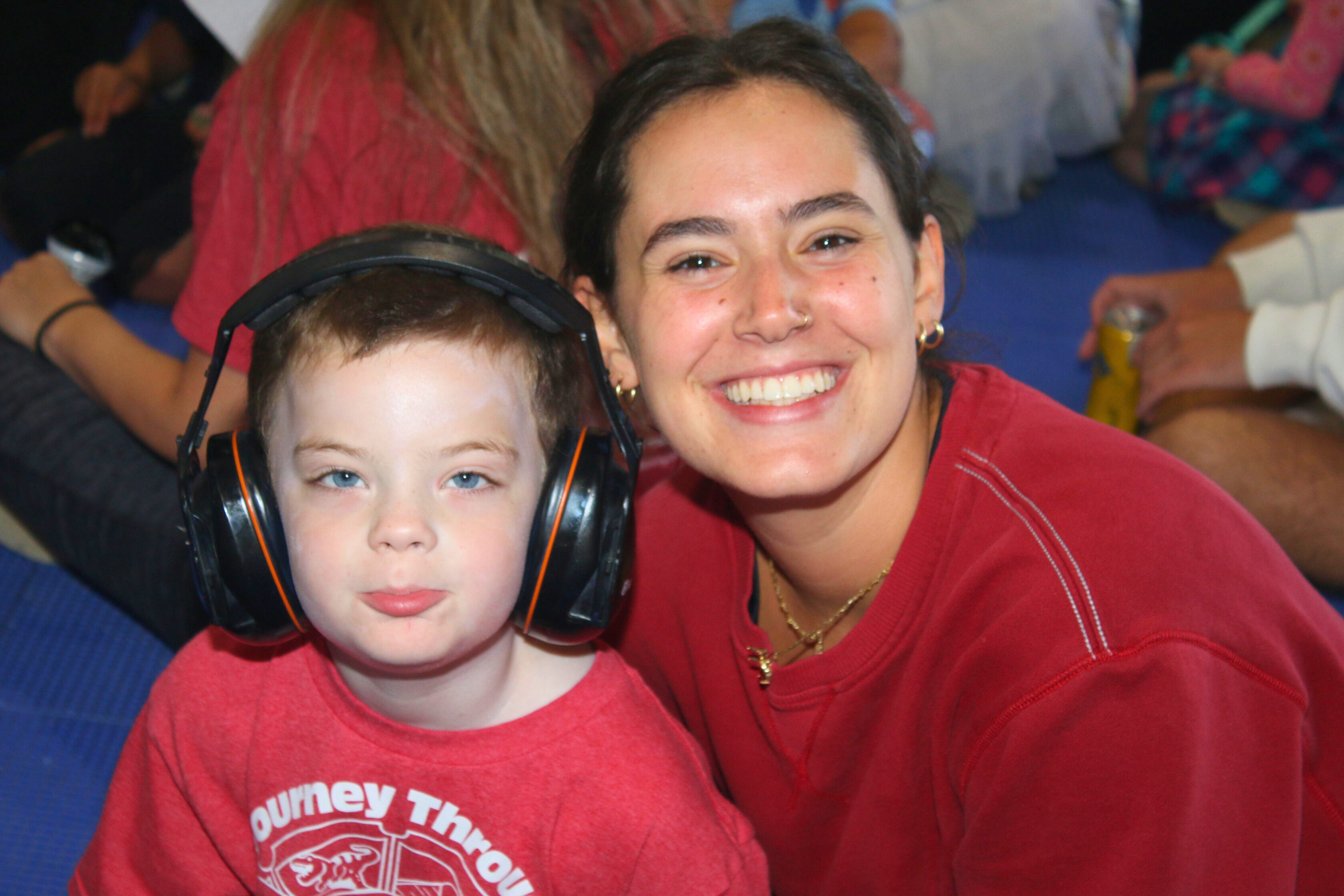 A young child wearing noise-canceling headphones sits next to a smiling woman. Both are dressed in red shirts and are indoors with other people in the background.
