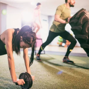 Three people exercise in a gym; one uses an ab roller, another flips a large tire, and the third jumps rope in the background.