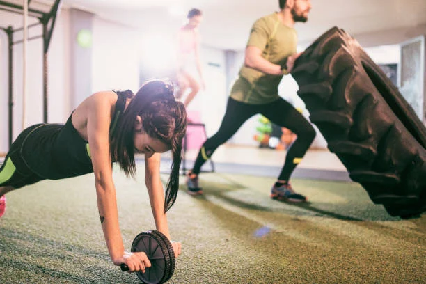 Three people exercise in a gym; one uses an ab roller, another flips a large tire, and the third jumps rope in the background.