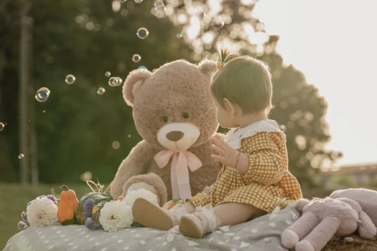 A baby in a yellow checked dress sits on a blanket outdoors, facing a large teddy bear, with bubbles floating nearby and flowers around them.