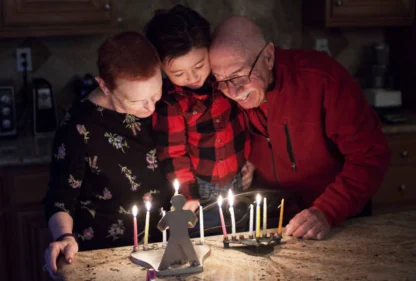 Two older adults and a child stand together, smiling and lighting Hanukkah menorahs with lit candles on a kitchen counter.