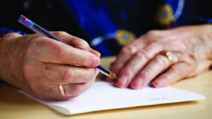 Close-up of an elderly person's hands writing with a pen on a piece of paper, wearing gold rings on both ring fingers.