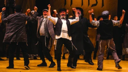 A group of men in traditional Jewish attire perform an energetic dance on stage, with raised arms and expressive faces.