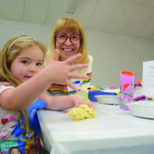 A young girl and an older woman sit at a table with craft supplies. The girl shows her hand to the camera while smiling.