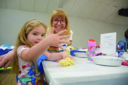A young girl and an older woman sit at a table with craft supplies. The girl shows her hand to the camera while smiling.
