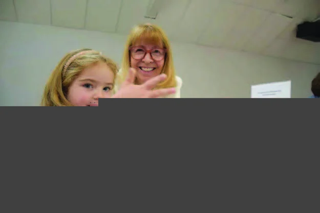 A young girl and an older woman sit at a table with craft supplies. The girl shows her hand to the camera while smiling.