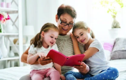 An older woman sits on a bed reading a book to two young girls, who are sitting close beside her and listening attentively.