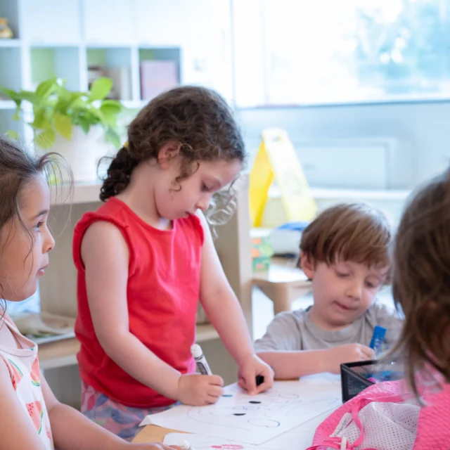 Four young children sit and stand around a table, drawing on paper with markers in a brightly lit classroom.