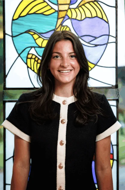 A woman with long brown hair stands smiling in front of a colorful stained glass window, wearing a black dress with white trim and gold buttons.
