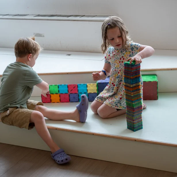 Two young children sit on steps indoors, building structures out of colorful magnetic tiles; one stacks blocks vertically while the other arranges them horizontally.