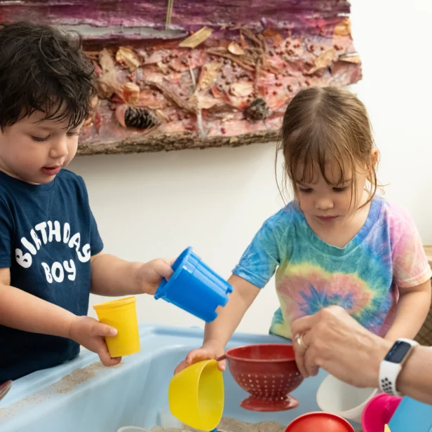Two young children play with colorful cups and containers at a sand table, supervised by an adult. A textured art piece hangs on the wall behind them.