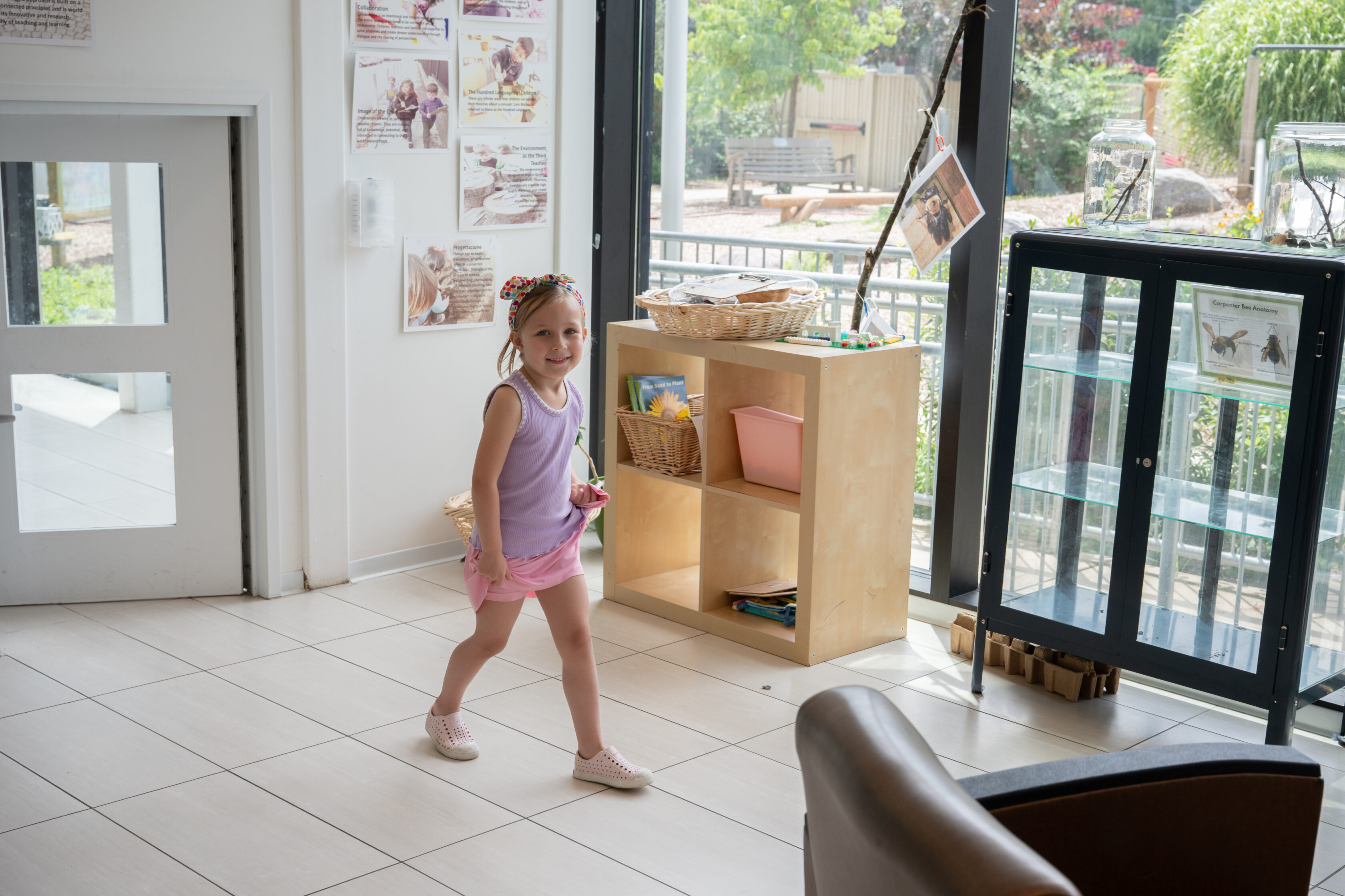 A young girl in a purple top and pink shorts walks and smiles inside a bright room with bookshelves and large windows looking out to a garden.