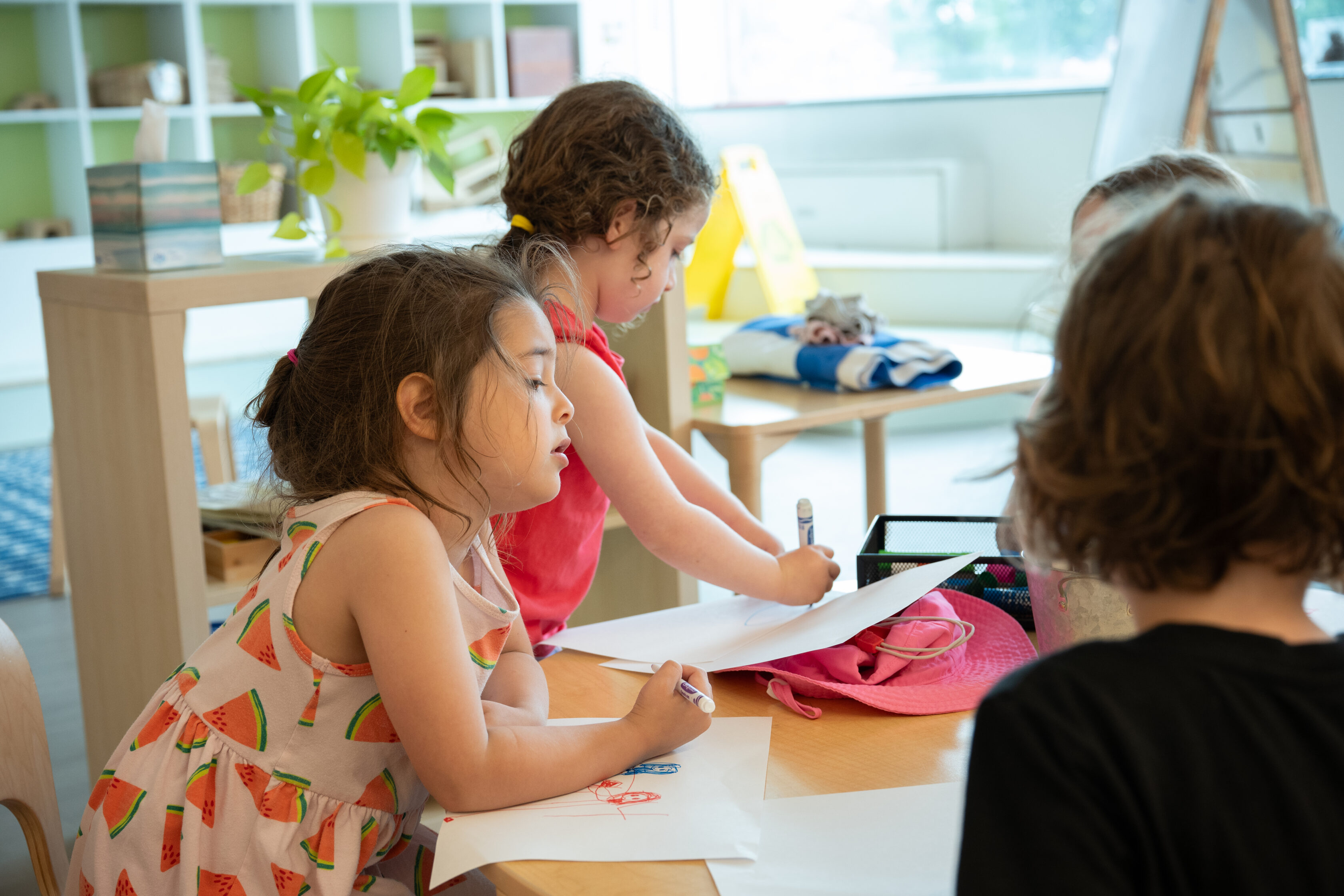Four young children are gathered around a table in a classroom, drawing on sheets of paper with markers.