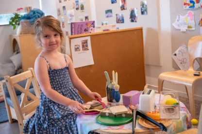 A young girl stands by a small table set with play food and utensils in a classroom decorated with children's artwork and photographs.